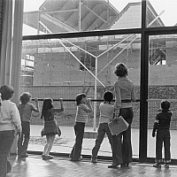 Black and white archival image of children and a teacher looking through large windows at a brick building under construction