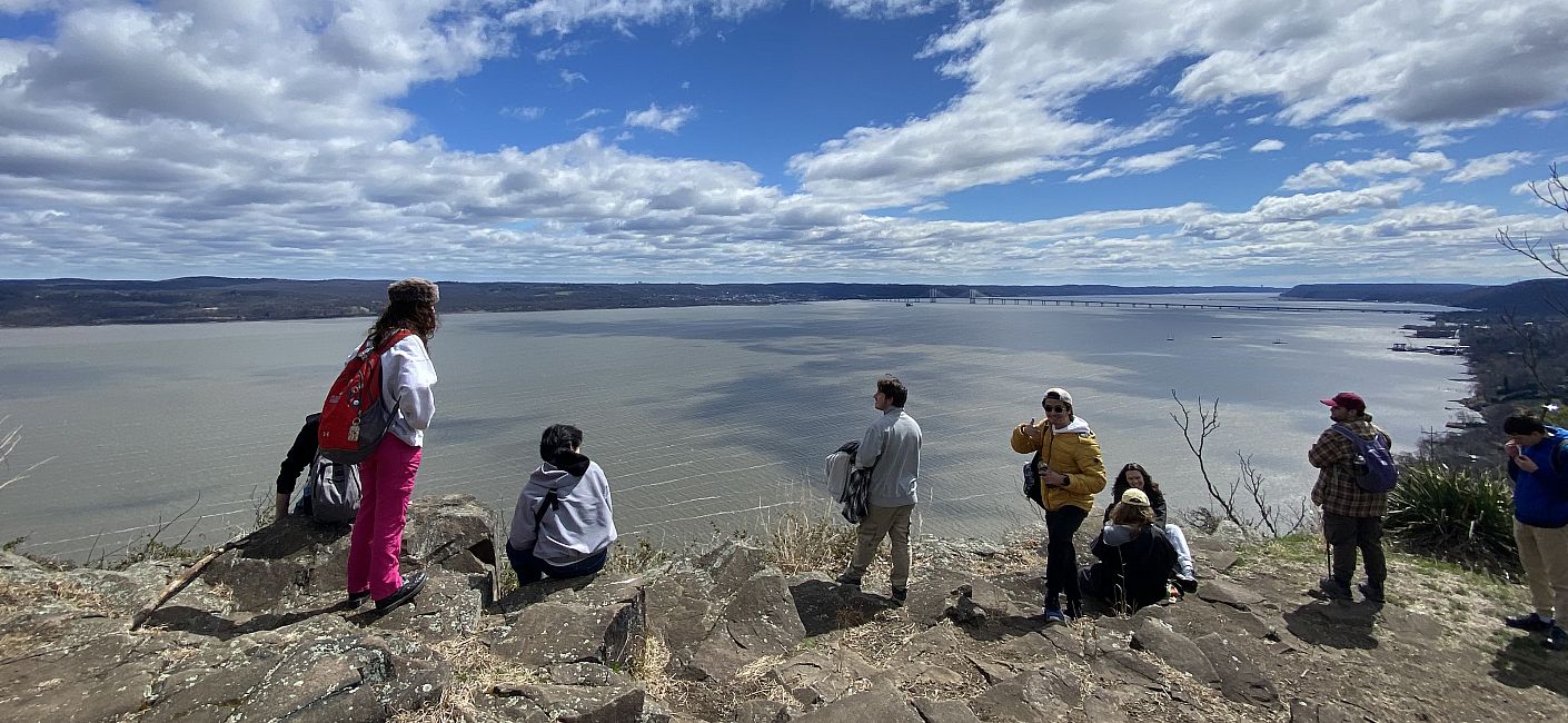 Several students stand scattered on a hilltop looking out at the  Hudson River in the distance.