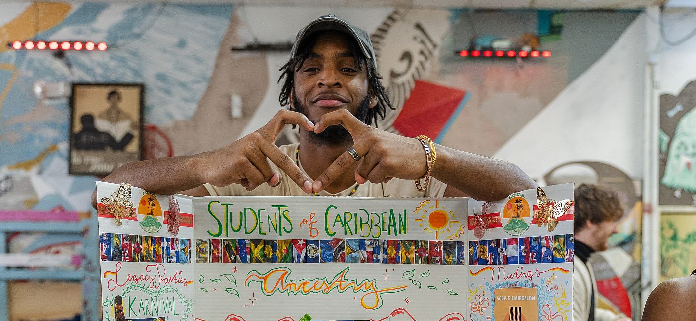 Student makes a heart with fingers standing behind a Students of Caribbean Ancestry Club sign.