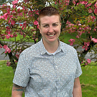 Alyssa is smiling standing in front of a tree with pink flowers. She has on a short sleeve collared white shirt with blue squares on it. 