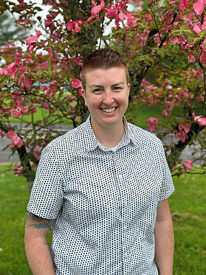 Alyssa is smiling standing in front of a tree with pink flowers. She has on a short sleeve collared white shirt with blue squares on it. 