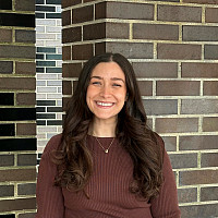 Gabriella is smiling in front of a brick wall. She has long brown hair and blue eyes. She is wearing a brown long-sleeved shirt.