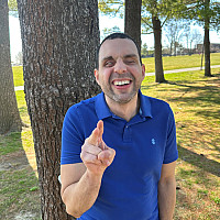 Ruben is wearing a blue, short-sleeve, collared shirt. He is standing in front of some trees with a big smile on his face, holding up the pe