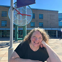 Tracey is smiling in front of the clock tower and the Student Services building. She is wearing a black shirt and has strawberry blonde, cur