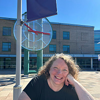 Tracey is smiling in front of the clock tower and the Student Services building. She is wearing a black shirt and has strawberry blonde, ...