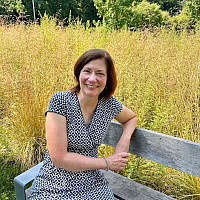 Kathy is sitting on a bench smiling. She is wearing a dress and there are wild yellow plants in the background.