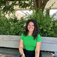 Gianna is smiling and sitting on a bench with a bright green shirt on. Her hair is dark and curly and falls to her shoulders.