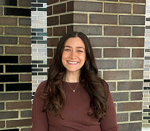 Gabriella is smiling in front of a brick wall. She has long brown hair and blue eyes. She is wearing a brown long-sleeved shirt.