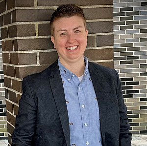 Alyssa is wearing a blue shirt with a blazer and is standing against a brick wall smiling.