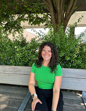 Gianna is smiling and sitting on a bench with a bright green shirt on. Her hair is dark and curly and falls to her shoulders.