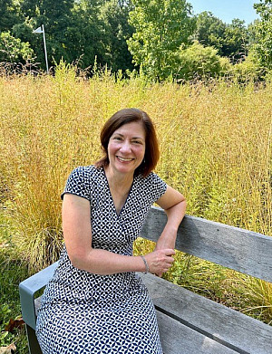 Kathy is sitting on a bench smiling. She is wearing a dress and there are wild yellow plants in the background.