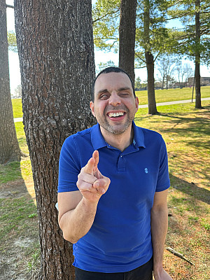 Ruben is wearing a blue, short-sleeve, collared shirt. He is standing in front of some trees with a big smile on his face, holding up the pe