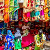 Colorful saris hang from the ceiling and walls of a market stall while customers shop.