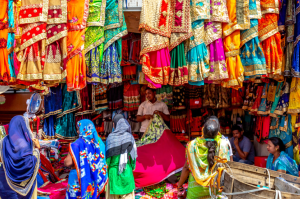 Colorful saris hang from the ceiling and walls of a market stall while customers shop.