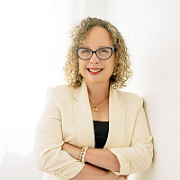 A woman with curl blond hair, wearing a white blazer, stands in front of a white background smiling with her arms crossed.