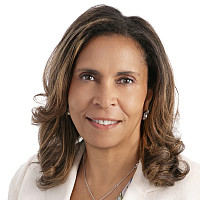 Woman with brown curly hair, wearing a white blazer with a floral top