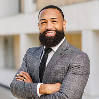 A man with a beard wearing a grey plaid suit jacket and black tie with his arms crossed over his chest.