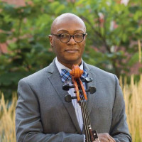 Earnest Lamb in gray suit, blue bow tie standing in front of tall grass background holding a cello