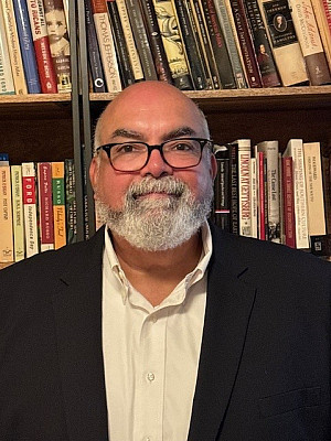 A man with a white beard wearing a white dress shirt and black jacket standing in front of a bookshelf