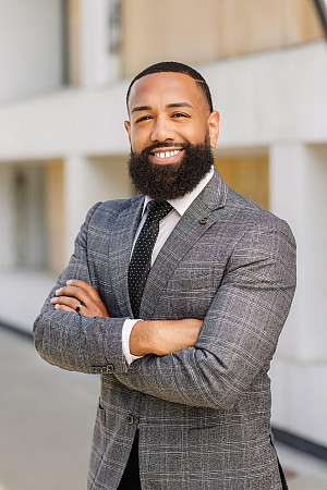 A man with a beard wearing a grey plaid suit jacket and black tie with his arms crossed over his chest.