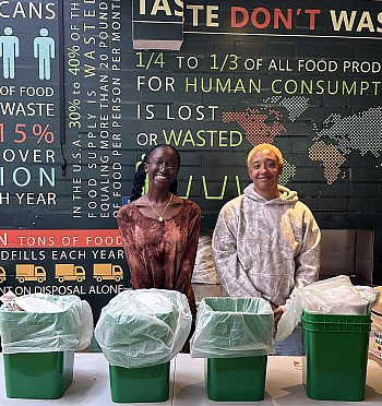 two students standing behind a table that has 4 green compost pails