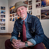 A man wearing a textured hat, denim jacket, red tie, and beaded bracelet sits smiling in front of a gallery wall filled with colorful photos