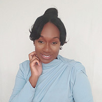 A woman with dark hair styled up, wearing a light blue long-sleeve top, sits against a plain white background, smiling gently.