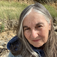 A woman with long gray hair and a blue jacket takes a selfie outdoors, with tall grass, rocks, and earth visible in the background under nat