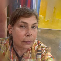 A woman with short brown hair and a floral shirt takes a selfie indoors. The background features bright yellow walls and a polished concrete