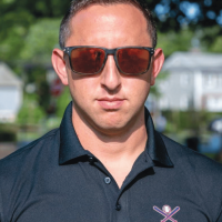 A man wearing sunglasses and a dark polo shirt with a crossed bats logo stands outdoors on a sunny day, with houses and greenery blurred in
