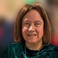A woman with straight brown hair wears a green velvet top and a gold cross necklace, smiling at the camera. The background is softly blurred