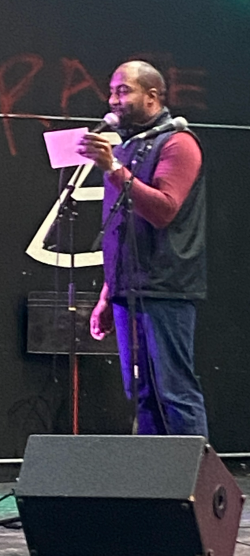 A man stands on stage holding a sheet of paper and speaking into a microphone. He is wearing a maroon long-sleeve shirt, jeans, and a black