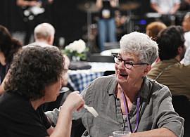 Two women sit at a table eating and laughing together at an indoor event, while a live band performs on stage in the background. Other guest