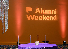 Projected image of Alumni Weekend on a yellow wall with event decor on the tables and suspended from the ceiling