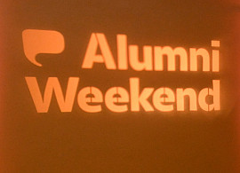 Projected image of Alumni Weekend on a yellow wall with event decor on the tables and suspended from the ceiling