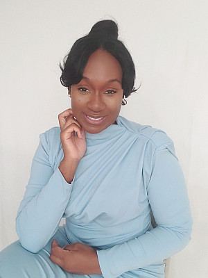 A woman with dark hair styled up, wearing a light blue long-sleeve top, sits against a plain white background, smiling gently.