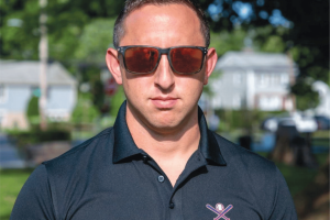 A man wearing sunglasses and a dark polo shirt with a crossed bats logo stands outdoors on a sunny day, with houses and greenery blurred in