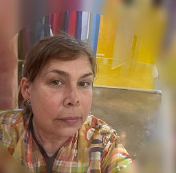 A woman with short brown hair and a floral shirt takes a selfie indoors. The background features bright yellow walls and a polished concrete