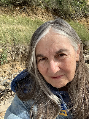 A woman with long gray hair and a blue jacket takes a selfie outdoors, with tall grass, rocks, and earth visible in the background under nat