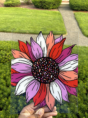 A hand holds up a piece of clear glass painted with a vibrant, stylized sunflower in shades of red, pink, orange, and white, with green bu