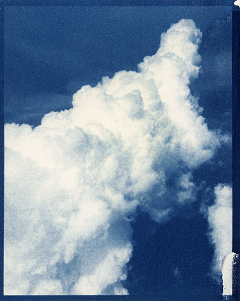A large, fluffy white cloud stretches across a deep blue sky, captured in a cyanotype style that adds a vintage, blue-tinted effect to the i
