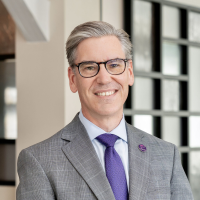 A man with gray hair and glasses, wearing a pink suit jacket, white shirt, and checkered tie, smiles while standing outdoors.