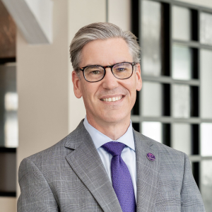 A man with gray hair and glasses, wearing a pink suit jacket, white shirt, and checkered tie, smiles while standing outdoors.