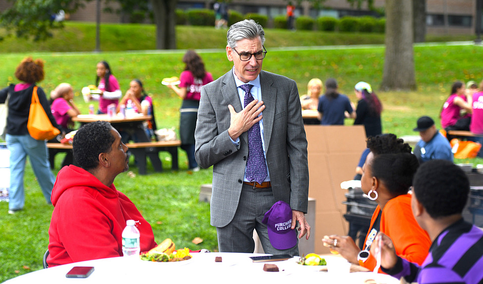 Man in suit stands speaking to adults sitting at a table turned toward him.