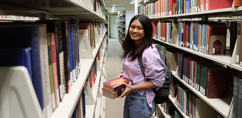 Female student in pink sweatshirt smiles standing in library stacks