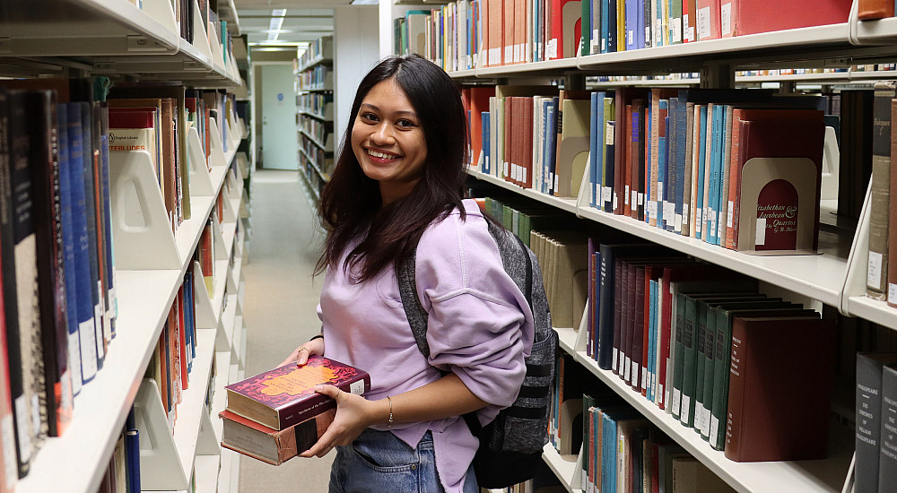 Female student in pink sweatshirt smiles standing in library stacks