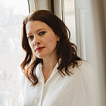 Color photograph of a woman with long brown hair wearing a white blouse standing in front of a window