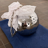 A woman is reflected in a shiny metal sphere with small holes sits on a blue padded mat. A crumpled paper protects the surface.