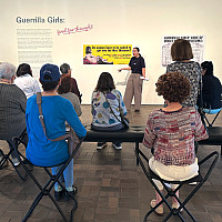 A group of museum visitors sits facing a wall with artwork, listening to a guided tour