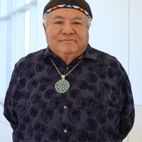 An older man stands indoors wearing a patterned dark shirt, a beaded cap, and a large turquoise necklace, with his hands behind his back and